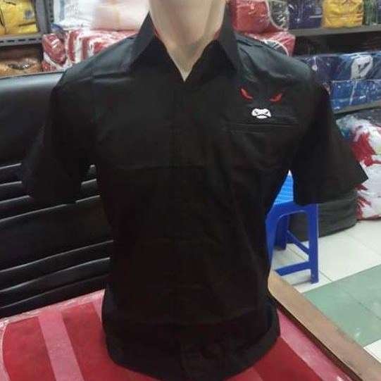 Kemeja hitam lengan pendek dengan logo hidung banteng mata merah dan mulut marah di saku dada kiri, dipajang di atas manekin di dalam toko pakaian.